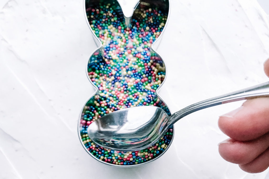 Spoon pushing sprinkles down inside a bunny cookie cutter on iced cake